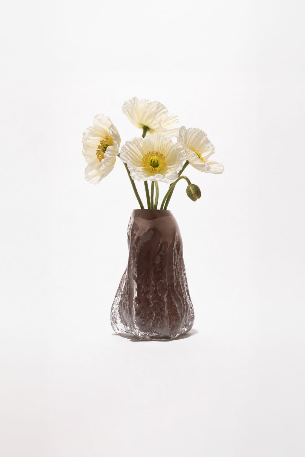 A smoky cast-glass vase holding white poppies, on white